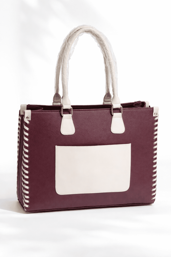A structured burgundy handbag with a rectangular shape, featuring cream-colored double handles and matching trim. The bag has a large cream front pocket and decorative side stitching, giving it a clean, modern look.